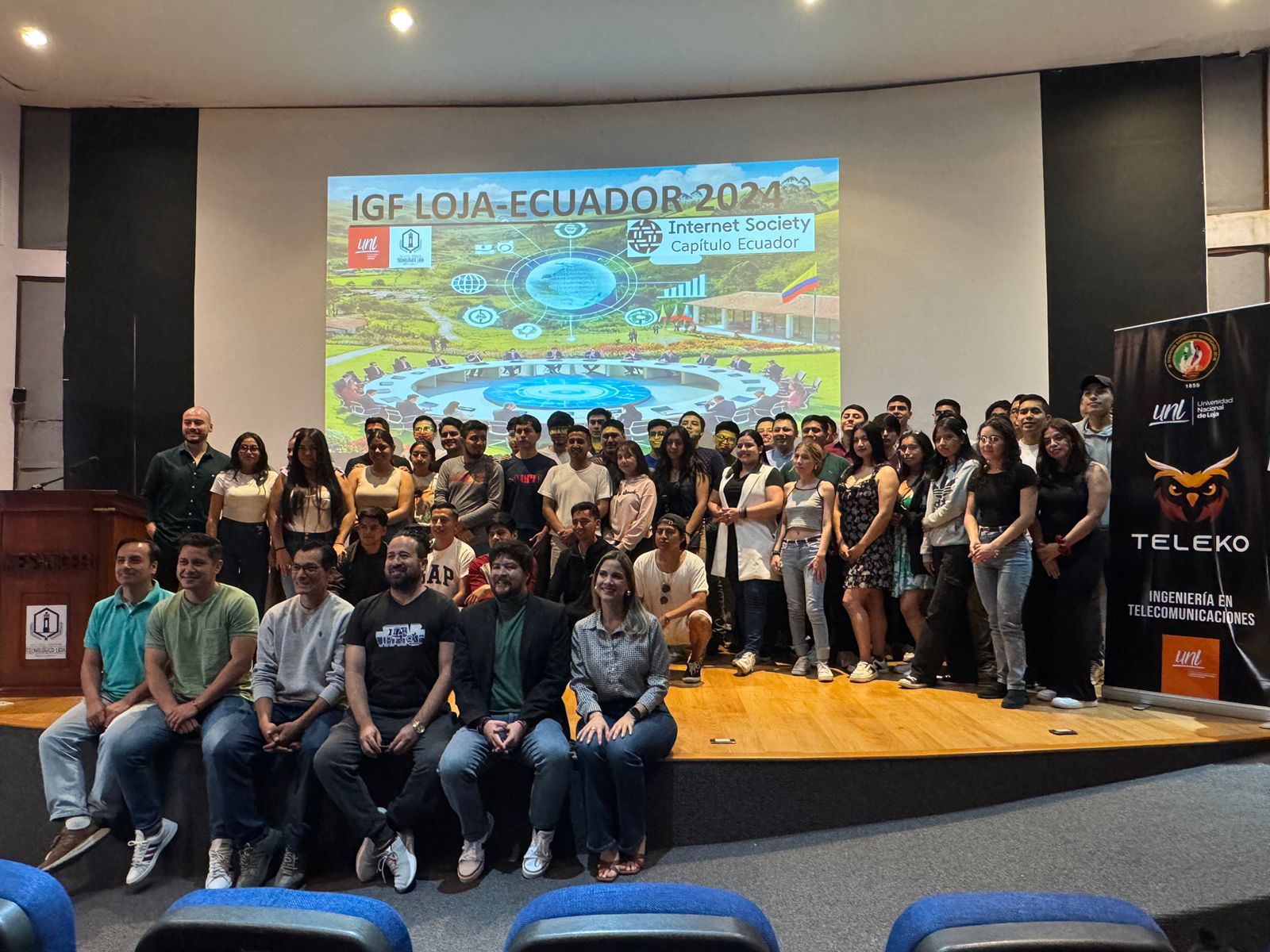 Grupo de estudiantes y organizadores en el escenario de un encuentro de gobernanza de Internet, con el logo de Internet Society Capítulo Ecuador proyectado al fondo